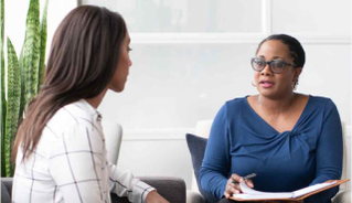 Two women sit across from each other in conversation, one appearing to listen attentively while holding a pen and clipboard.