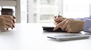 Hands facing eachother on a desk holding coffee and writing utensil, respectively