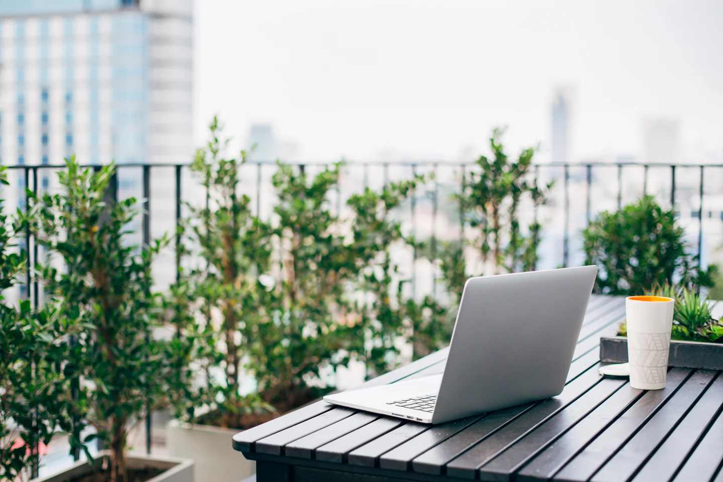 An open laptop on a patio table with plants and a railing.