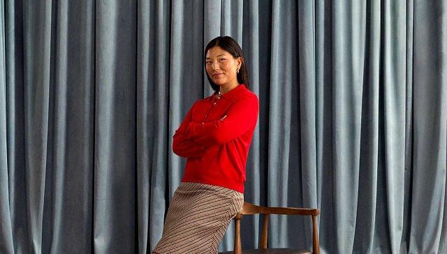 woman wearing a red shirt and beige skirt leaning on the side of a wooden chair