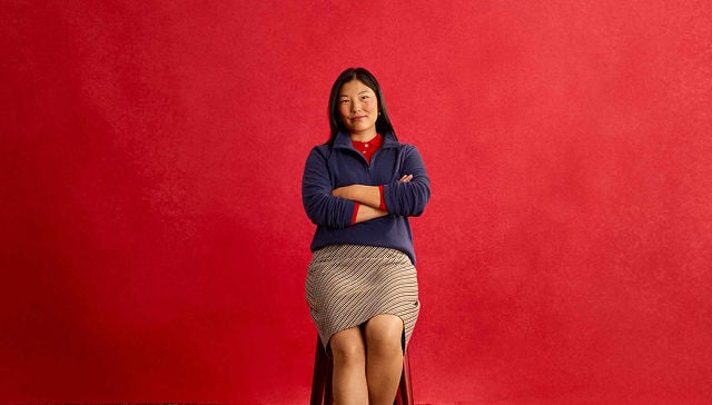 woman with a red shirt, blue sweater, and beige skirt sitting in a chair