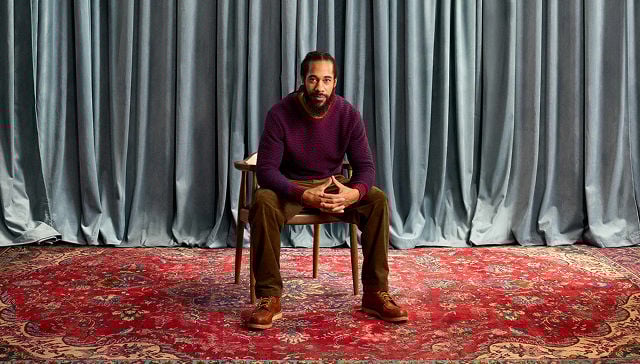 man in purple sweater, brown pants, and boots sitting in a chair in front of drapes