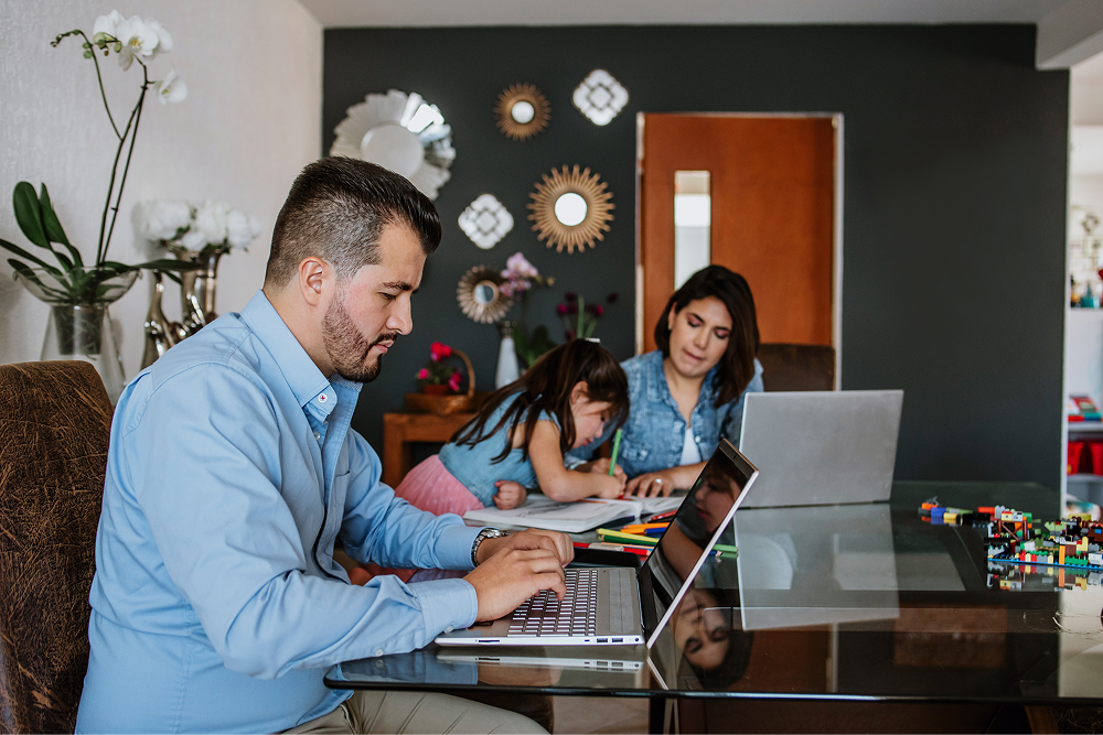 Portrayal of Capella University student working on a laptop at home with family in the background.