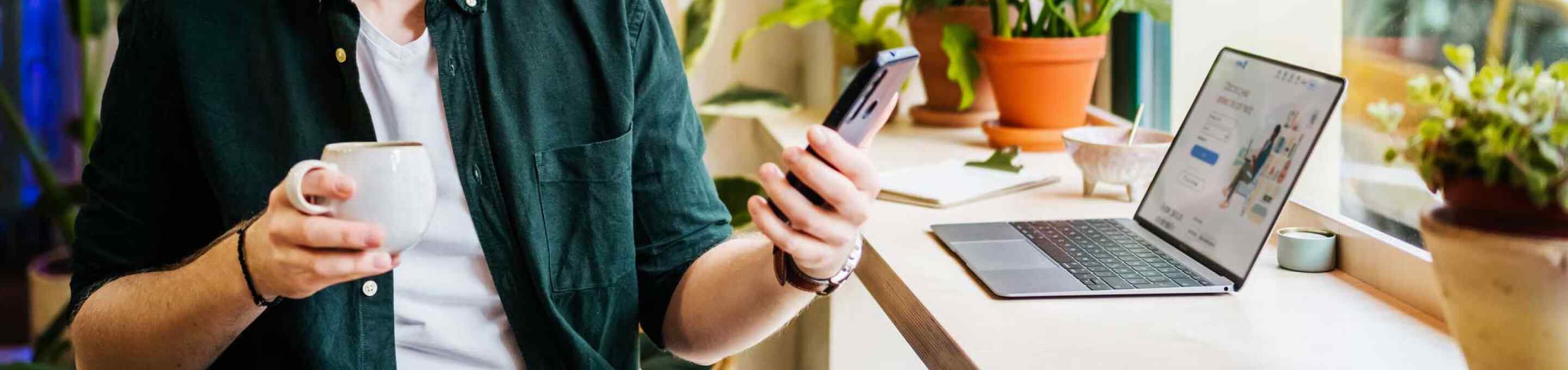 man in green shirt holding a phone and a cup of coffee, next to a laptop