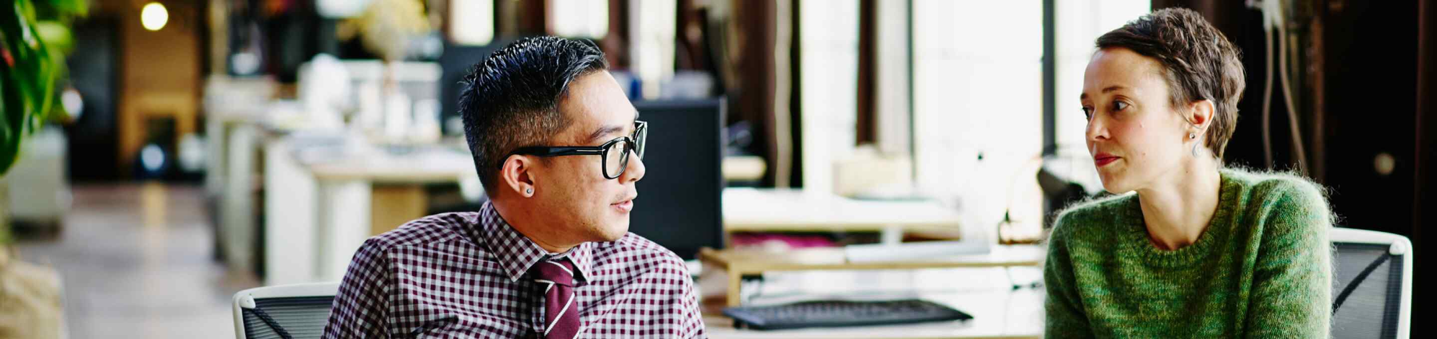 man in a red plaid shirt and tie talking to a woman in a green sweater