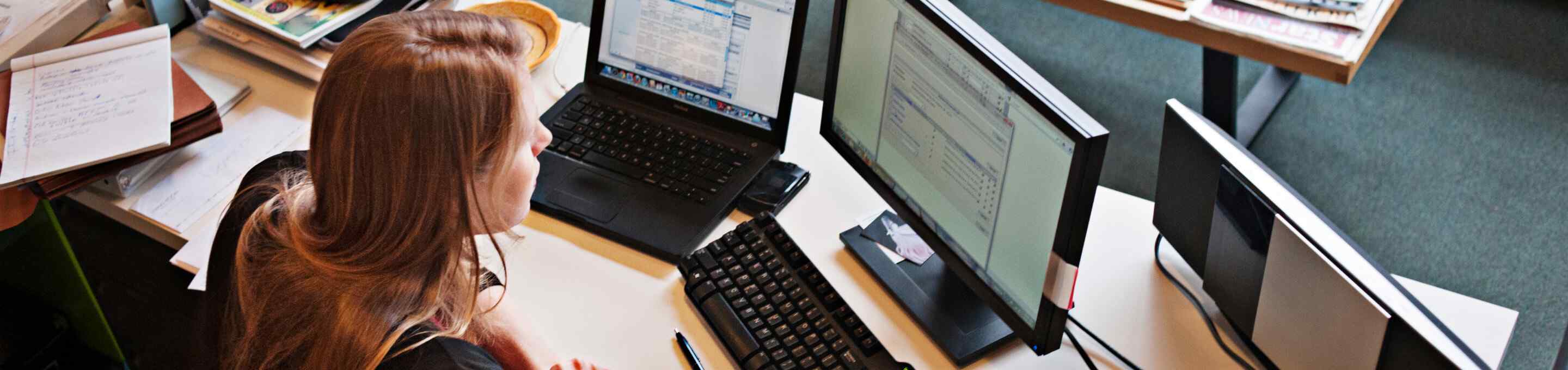 woman with red hair working while looking at two monitors