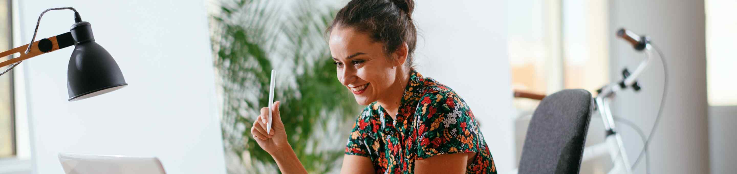 woman in floral shirt holding a pen while smiling at a computer screen