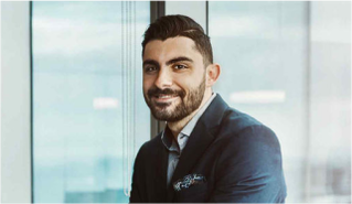 Smiling man with dark hair and a beard wearing a navy blazer, standing in front of a large window in a modern office.
