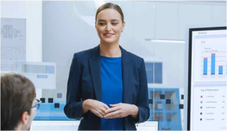 A businesswoman in a navy blazer and blue top presents data to a colleague in a modern office, with charts displayed on a nearby screen.
