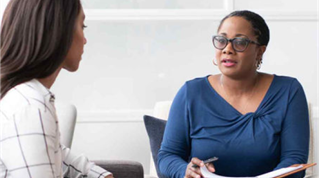 Counselor woman speaking to a client across from her, holding a pen and notepad in a bright office setting