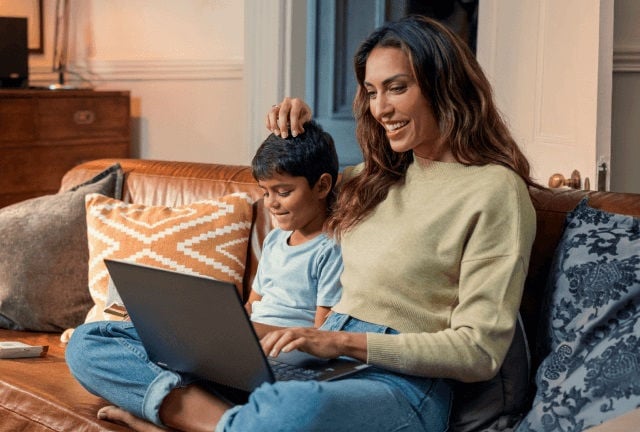 A woman and a young boy sitting together on a leather couch, smiling as they look at a laptop on her lap in a cozy living room.
