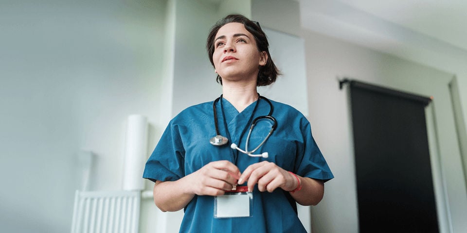 A female nurse in blue scrubs stands upright in a softly lit room, focused as she looks ahead, holding a lanyard.